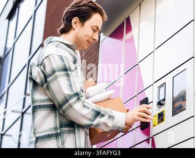 Man using smartphone at automated parcel locker for contactless delivery pickup. Modern self-service terminal for online shopping logistics and courie Stock Photo