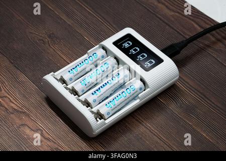 Set of four AAA rechargeable batteries in a white electronic charger with an LCD status display on a dark wood background. Stock Photo