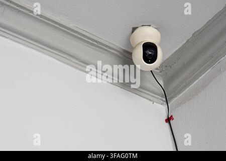 Modern white spherical security camera installed on an indoor ceiling corner for safety and property monitoring. Features a black power cable against Stock Photo