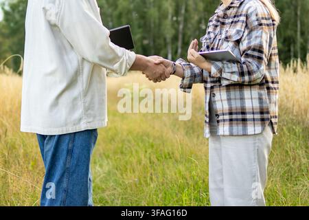 Faceless agricultural partners shake hands in wheat field while holding tablets together. Concept of anonymous business agreement and agricultural col Stock Photo