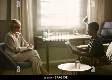 Middle aged Caucasian female therapist taking notes during session with young African American female soldier. Image suitable for mental health and military reintegration campaigns Stock Photo