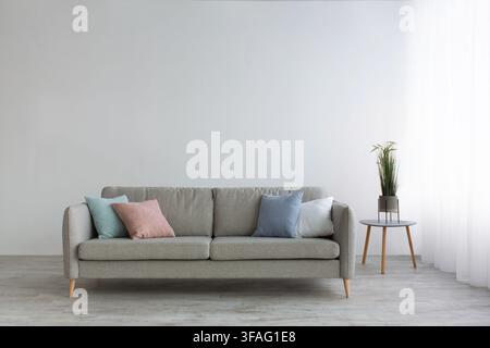 Gray sofa with pillows, table with plants and white curtains on window, on light wall background in living room Stock Photo