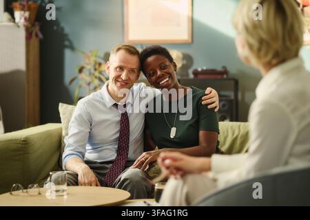 Happy Caucasian middle aged man and Black middle aged woman sitting on sofa during therapy session. Diverse couple smiling at counselor, ideal for mental health and relationship counseling ads Stock Photo