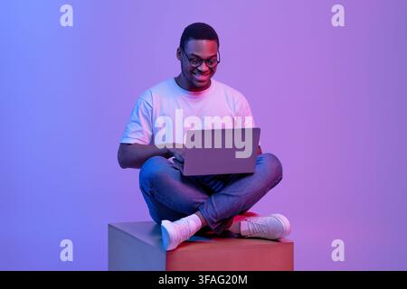 Black Programmer Guy Using Laptop Computer, Sitting On Cube In Neon Lighting Stock Photo
