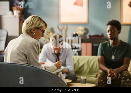 Middle aged Caucasian female therapist taking notes during counseling session with African American female soldier and partner, representing PTSD support and mental health services Stock Photo