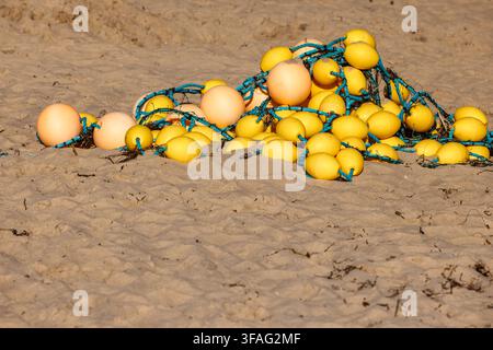 A close-up shot of a tangled pile of commercial fishing floats, bright yellow and orange buoys, and blue ropes left on fine sand. The beach surface is Stock Photo