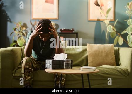 Stressed African American young female soldier sitting on sofa holding head in hands, suffering from PTSD. Image represents mental health support and therapy for veterans Stock Photo