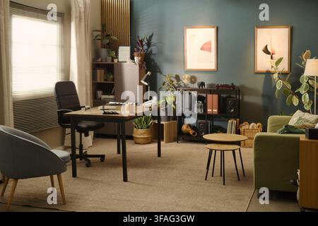 Empty modern home office interior showing desk with chair, shelving unit, and green sofa. Image suitable for remote work, hybrid workplace, and interior design marketing, wide shot Stock Photo