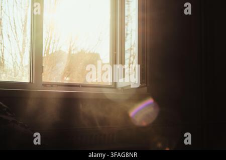 Bright morning sunlight streams through an open window into a room filled with swirling smoke and glowing dust particles illuminated by the warm light Stock Photo