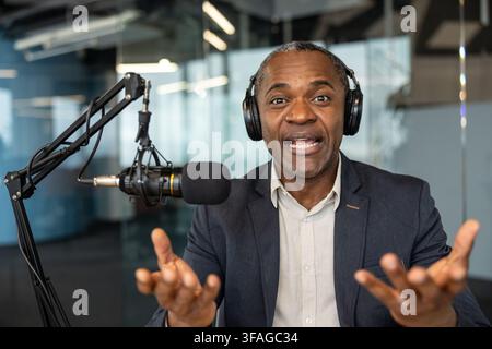 Mature man wearing headphones and speaking into a professional microphone, gesturing with his hands while recording a podcast or live streaming conten Stock Photo