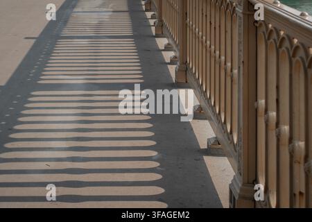 Ornate bridge railing casts long, rhythmic shadows across a sunlit pavement walkway, creating a striking pattern of light and shadow along the Stock Photo