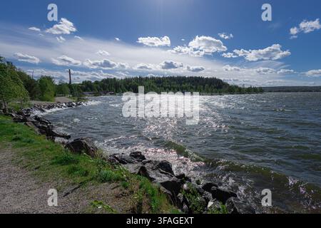 Sunlight sparkles across a wide, wind-rippled lake in Lahti, bordered by a rocky shoreline path and dense pine forest under a bright blue sky dotted Stock Photo