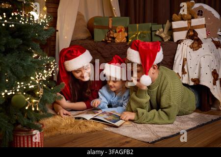 Three children wearing Santa hats, lying on a rug next to a glowing Christmas tree and reading a book together in a warmly decorated holiday interior Stock Photo