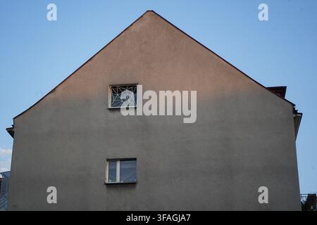 "Minimalist view of a triangular house wall with small windows against a clear blue sky, where one window features an intricate stained glass pattern. Stock Photo