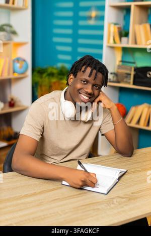African American young man on home table writes notes in notebook with pen for study planning at home. Black guy on home table reviews written task list with happy focused mood in living room interior Stock Photo