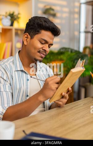 Indian man on home table reads book makes notes and pauses to think during focused study indoors at home. Arabian guy on home table reviews pages and writes ideas for learning planning and home work. Stock Photo