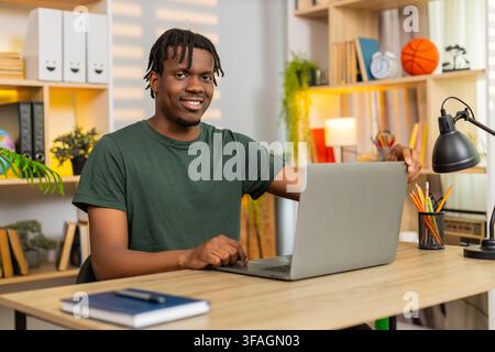 African American young man on home table opens laptop then starts focused remote work with calm smile in living room. Black guy on home table types email using computer for online job freelance task. Stock Photo