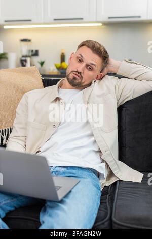 Young man at home works on laptop feels tired and demotivated and takes deep sigh during hard task. Caucasian guy on sofa slumps shoulders shows fatigue stress low energy and tries to focus now again Stock Photo