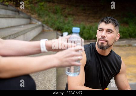 Sporty woman sitting on outdoor stairs and drinking bottled water while resting and hydrating after exercise. Stock Photo