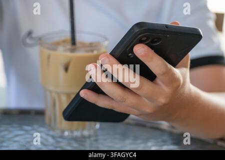 a person's hand holding and using a black smartphone, with a refreshing glass of iced coffee softly blurred in the background at a cafe table. Stock Photo