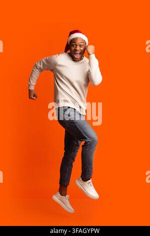 Ready For Party. Cheerful African Guy In Santa Hat Running In Air Stock Photo