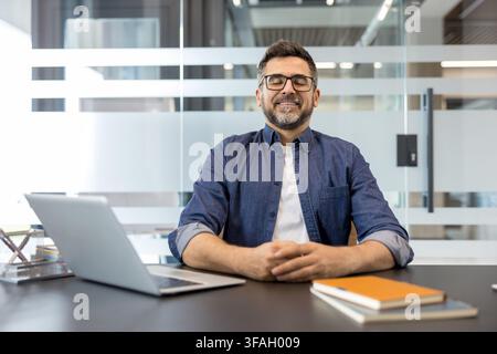 Middle-aged man with closed eyes and clasped hands sitting at an office desk with a laptop and notebooks, practicing mindfulness for stress relief and focus Stock Photo