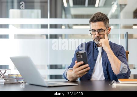 Confident businessman wearing glasses using his mobile phone and laptop at a desk in a modern office, concentrating on digital communication and online tasks Stock Photo