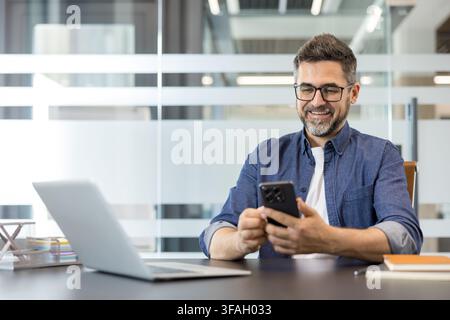 Businessman using a mobile phone and smiling while sitting at his desk with a laptop, communicating, networking, and browsing on his device in a modern corporate office setting Stock Photo