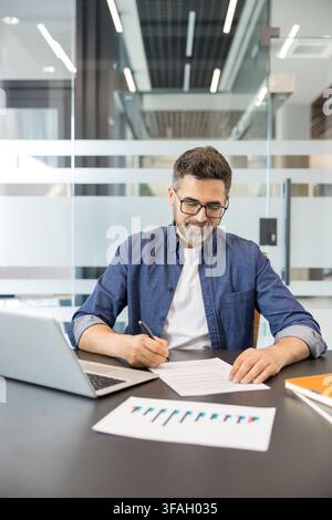 Professional businessman working in a modern office, reviewing paperwork, writing notes on a document, and analyzing business data on charts with a laptop nearby Stock Photo