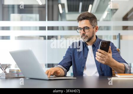Businessman concentrating on work. Simultaneously typing on a laptop and checking content on a smartphone. Representing efficiency. Digital communication Stock Photo