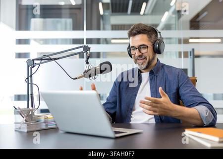 Mature man presenting and discussing a topic during a podcast recording session, communicating with listeners while sitting at his desk in a modern office Stock Photo