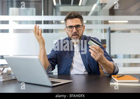 Frustrated man holding a credit card and shrugging, having problems with an online payment or e-commerce transaction while looking at a laptop in a modern office setting Stock Photo