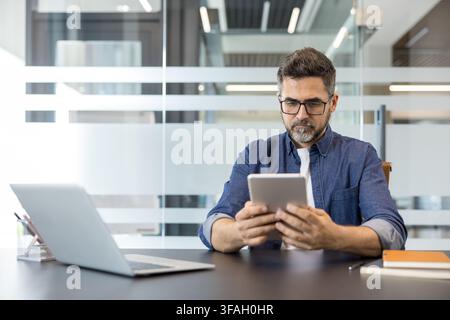 Mature man with glasses wearing a denim shirt, concentrating while using a digital tablet at his desk in a contemporary office setting, with a laptop and notebooks nearby Stock Photo