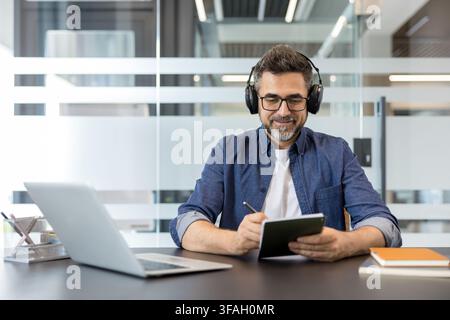 Mature man wearing headphones and glasses smiling, working productively at a modern office desk, making notes in a digital notebook with a stylus, and using a laptop Stock Photo