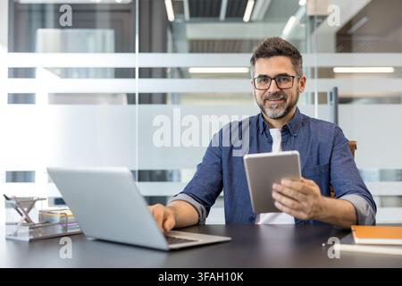 Confident man in a modern office using a laptop and a digital tablet, staying connected and productive while managing his business tasks with technology Stock Photo