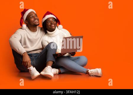 Dreamy african couple in santa hats sitting with laptop on orange background Stock Photo