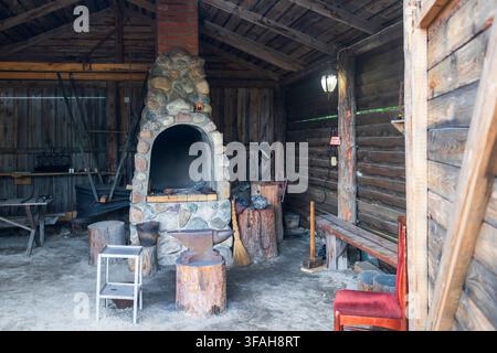 Traditional blacksmith workshop interior with stone-and-brick forge, iron anvil on log stump, hammer, broom, and various tools on earthen floor. Stock Photo