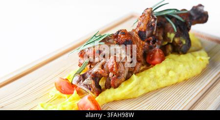 Roasted pork knuckle with crispy skin served on creamy mashed potato with cherry tomatoes and rosemary, on a wooden board. Hearty restaurant dish. Stock Photo