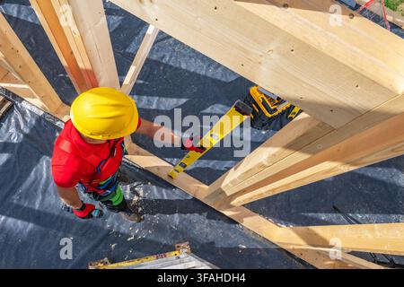 A construction worker in a yellow helmet is using a level tool to ensure the wood frame is straight at a building site. The worker is focused on their Stock Photo