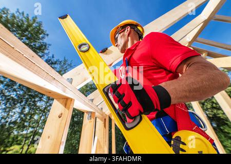 A worker holds a level tool while measuring wooden beams at a construction site. The location has bright skies and trees in the background. The worker Stock Photo
