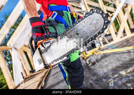 A construction worker stands on a building site while holding a chainsaw. The worker is wearing safety gear and prepared for wood cutting tasks. Surro Stock Photo