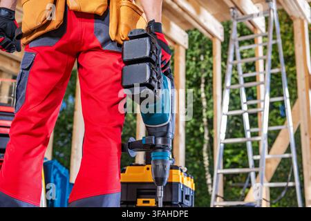 A worker stands at a construction site wearing a tool belt. They hold a power tool in one hand. In the background, there is a ladder and various tools Stock Photo