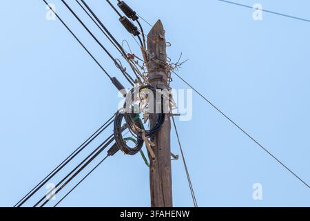 A close-up view of a messy bundle of black cables and wires coiled around an old, weathered wooden utility pole against a clear blue sky Stock Photo