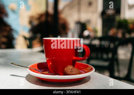Fresh hot coffee served in a red ceramic cup with cookie on a bright cafe table. Warm sunlight and modern coffee culture create a stylish lifestyle Stock Photo