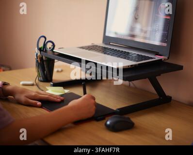 Digital artist drawing on a graphic tablet with a stylus at a wooden desk with a laptop on a stand in a cozy home office setup. Stock Photo