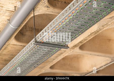 A detailed view of a modern industrial cable management system featuring a perforated metal tray suspended from a concrete ceiling structure Stock Photo