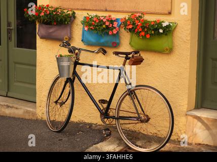 Vintage bicycle with metal bucket basket parked against yellow wall with colorful flower planters in street of Rijeka, Kvarner, Croatia. Stock Photo