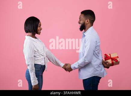 Loving black guy holding his sweetheart's hand, hiding present for her behind his back over pink background, side view Stock Photo
