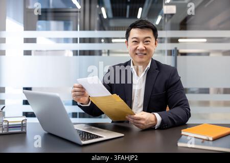 Asian businessman smiling and holding an open brown envelope, extracting important documents from it while sitting at his office desk with a laptop and notebooks Stock Photo