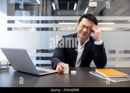Asian businessman suffering from a severe headache or migraine, feeling stressed and exhausted while trying to work on his laptop in a modern office, holding a bottle of pills Stock Photo